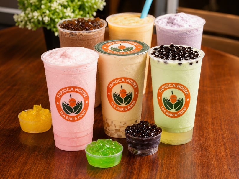 Six colorful Tapioca House bubble teas with boba and jellies on a wooden surface.