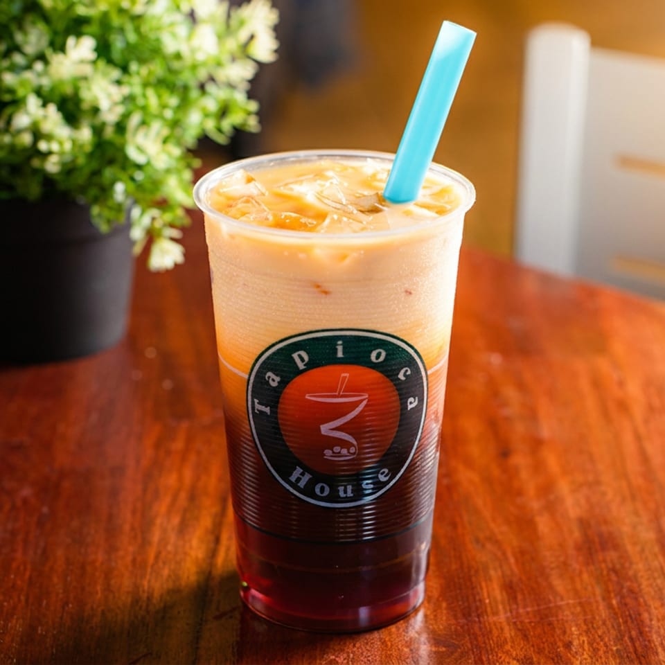 Layered Tapioca House drink with a blue straw on a wooden table next to a potted plant.
