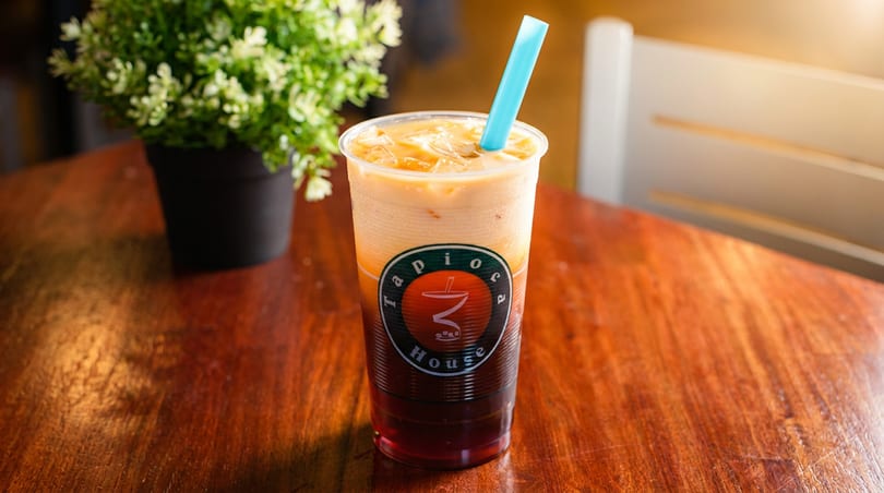 Layered Tapioca House drink with a blue straw on a wooden table next to a potted plant.