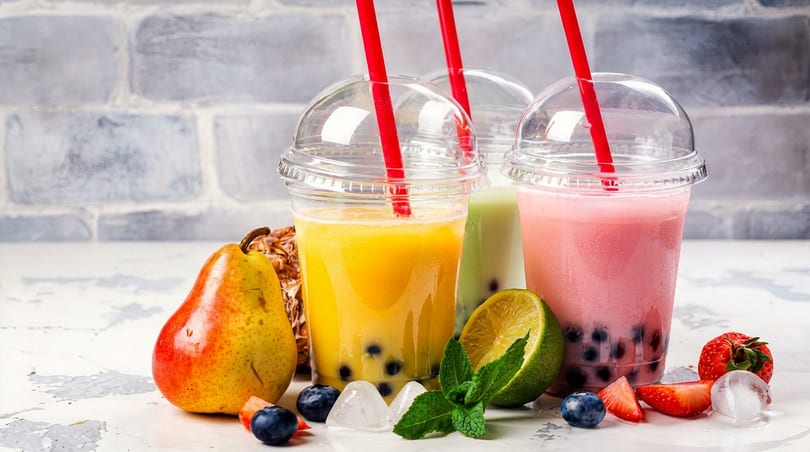 Assorted bubble teas in clear cups with red straws, fresh fruit, and ice cubes.