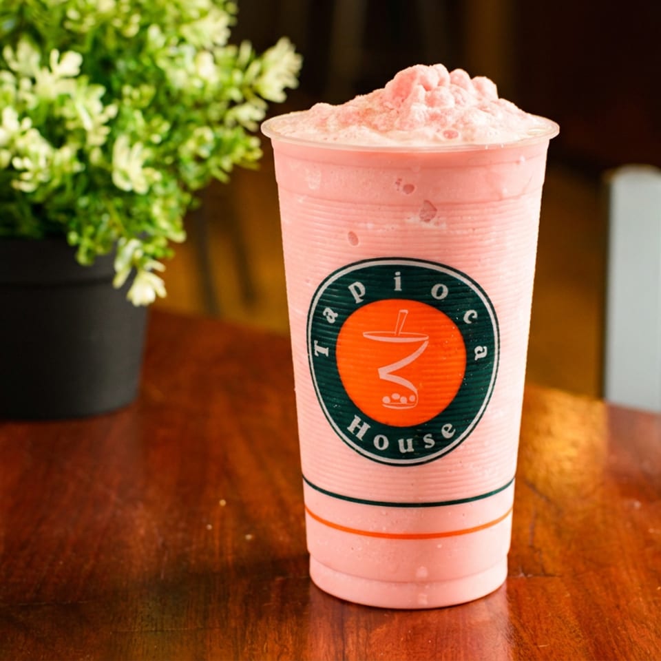Pink Tapioca House smoothie in a branded cup on a wooden table, next to a potted plant.