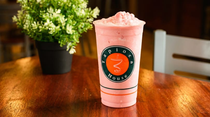Pink Tapioca House smoothie in a branded cup on a wooden table, next to a potted plant.
