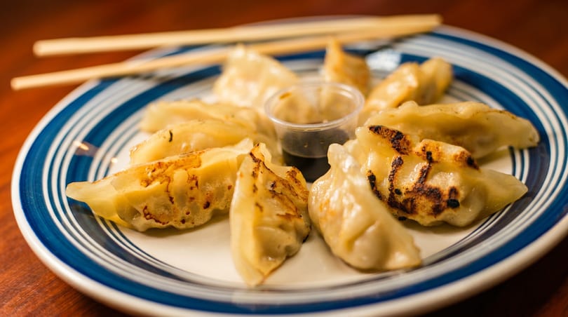 A plate of golden pan-fried dumplings with dipping sauce and chopsticks.